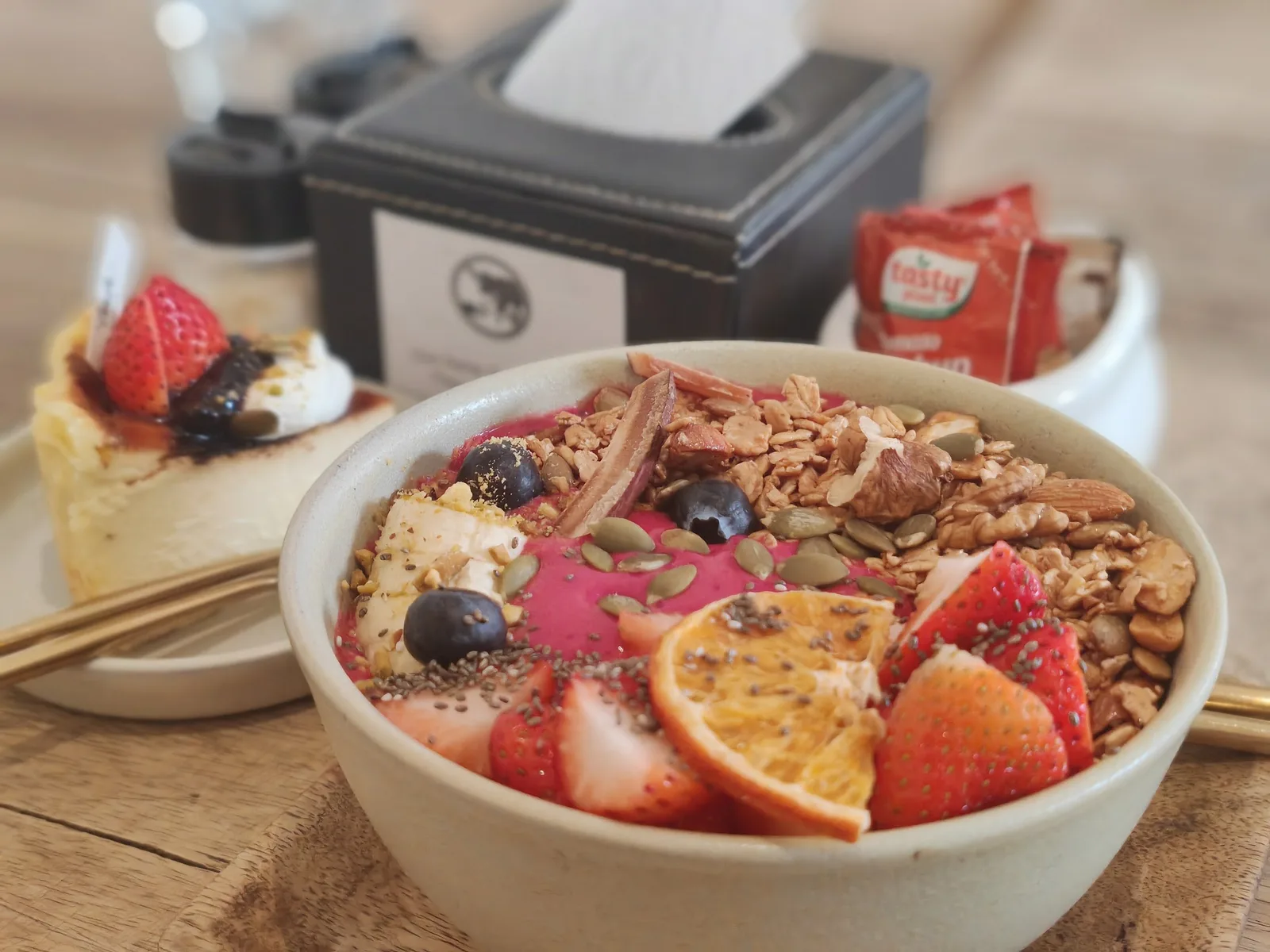 A slice of strawberry-topped cheesecake next to an açaí bowl on a wooden table at Cafe Camellia